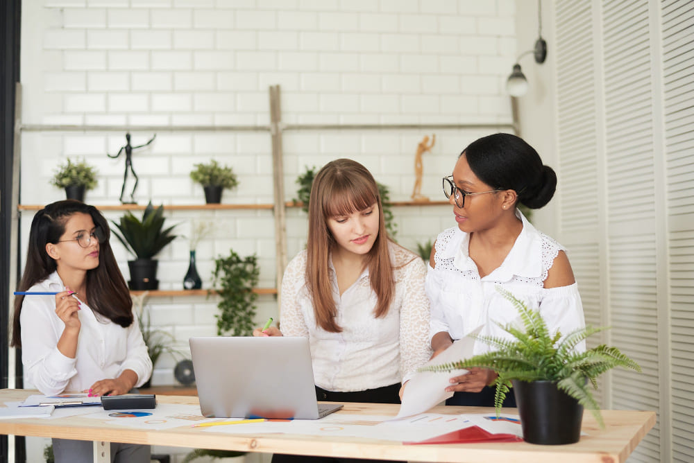 Serious female multiracial team solving problem together, diverse focused african, asian and caucasian colleagues discuss project brainstorming working on laptop, creative office teamwork.